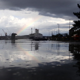 a ship partly framed by a rainbow, also reflected on the wet asphalt in the foreground