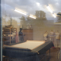 the inside of some kind of mystery tailor's workshop (?) photographed from the outside, through a window that's neither clean nor reflection-free. A black mannequin that looks like it's gesturing is casually leaning against one of the walls