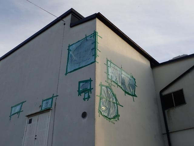 the corner of a building whose plaster is falling apart; each spot where it has come off is covered in a thin layer of blue semi-transparent plastic, held in place by some dark green tape