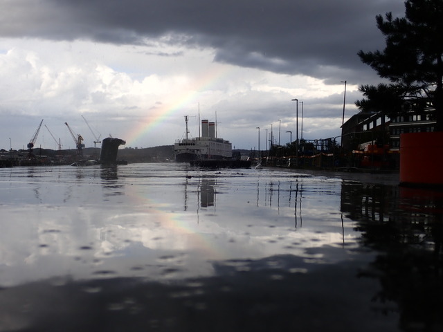 a ship partly framed by a rainbow, also reflected on the wet asphalt in the foreground