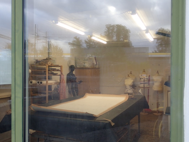 the inside of some kind of mystery tailor's workshop (?) photographed from the outside, through a window that's neither clean nor reflection-free. A black mannequin that looks like it's gesturing is casually leaning against one of the walls